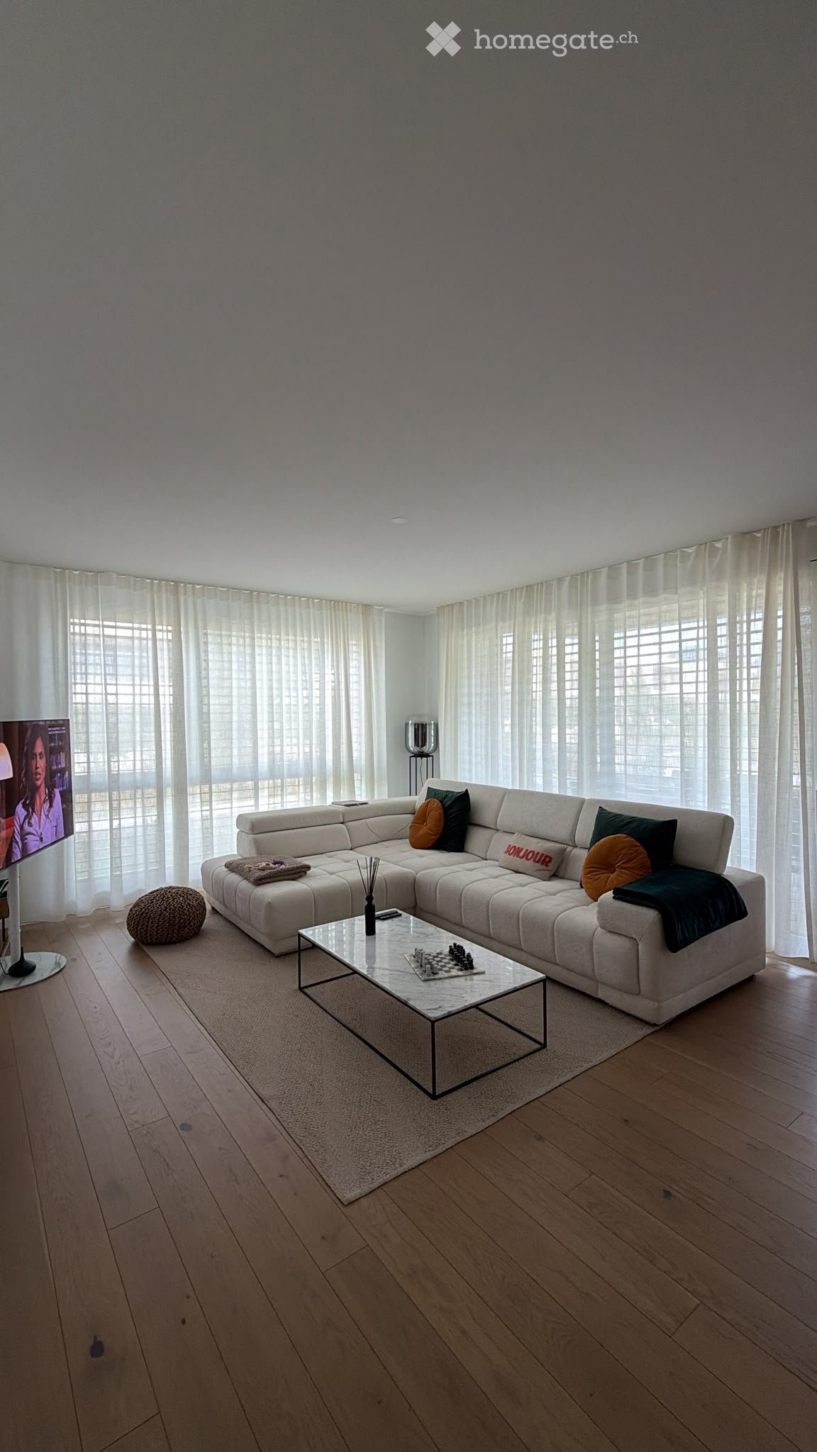 Modern living room with a large sectional sofa, coffee table, chessboard, floor lamp, and decorative ball. Large windows with curtains allow natural light to fill the space.