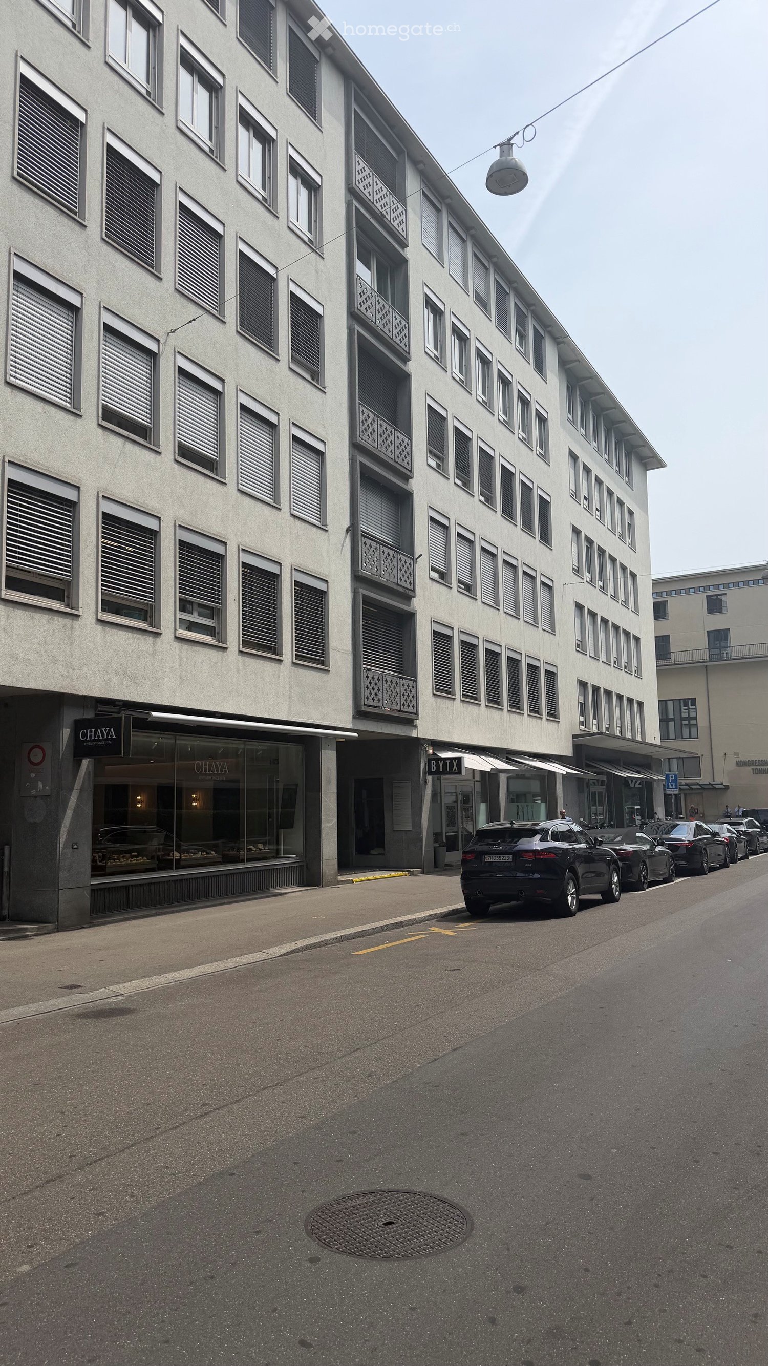 Multi-story building with numerous windows and balconies, located on a street with parked cars and storefronts.