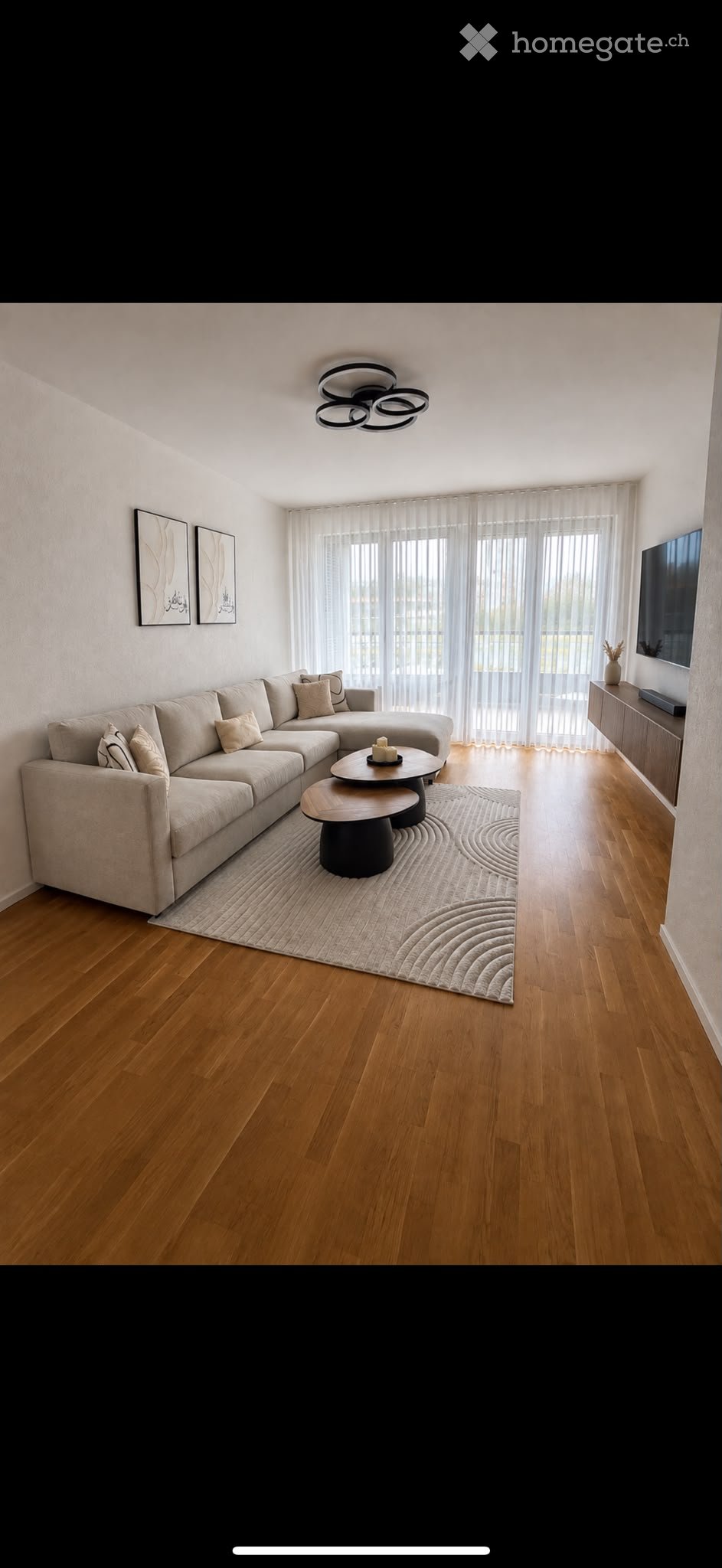 Modern living room with a sectional sofa, coffee table, TV, and large windows with sheer curtains.
