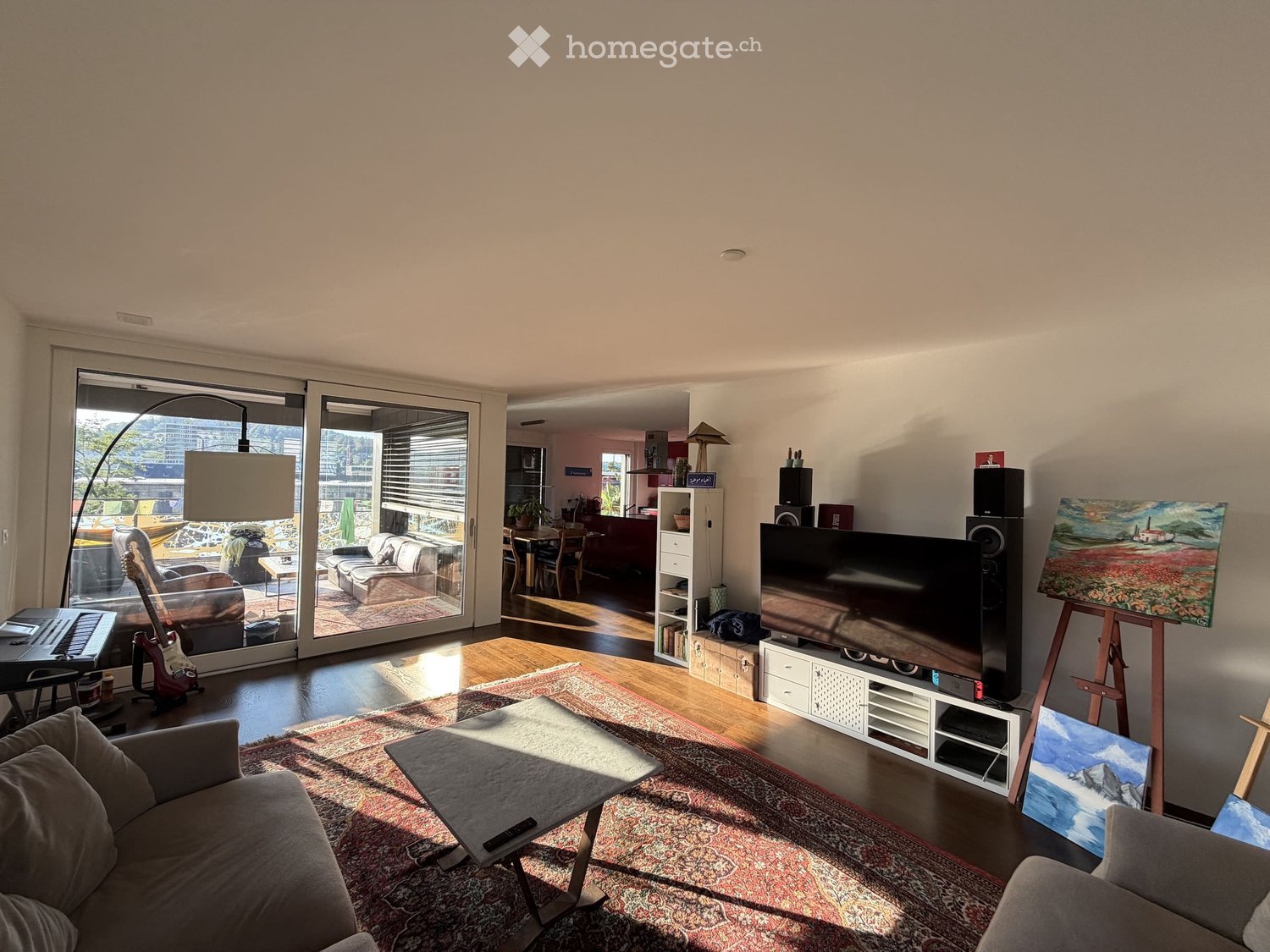 Bright living room with large sliding glass doors leading to a balcony, featuring a grey sofa, coffee table, TV, guitar, keyboard, floor lamp, and artwork on easels.