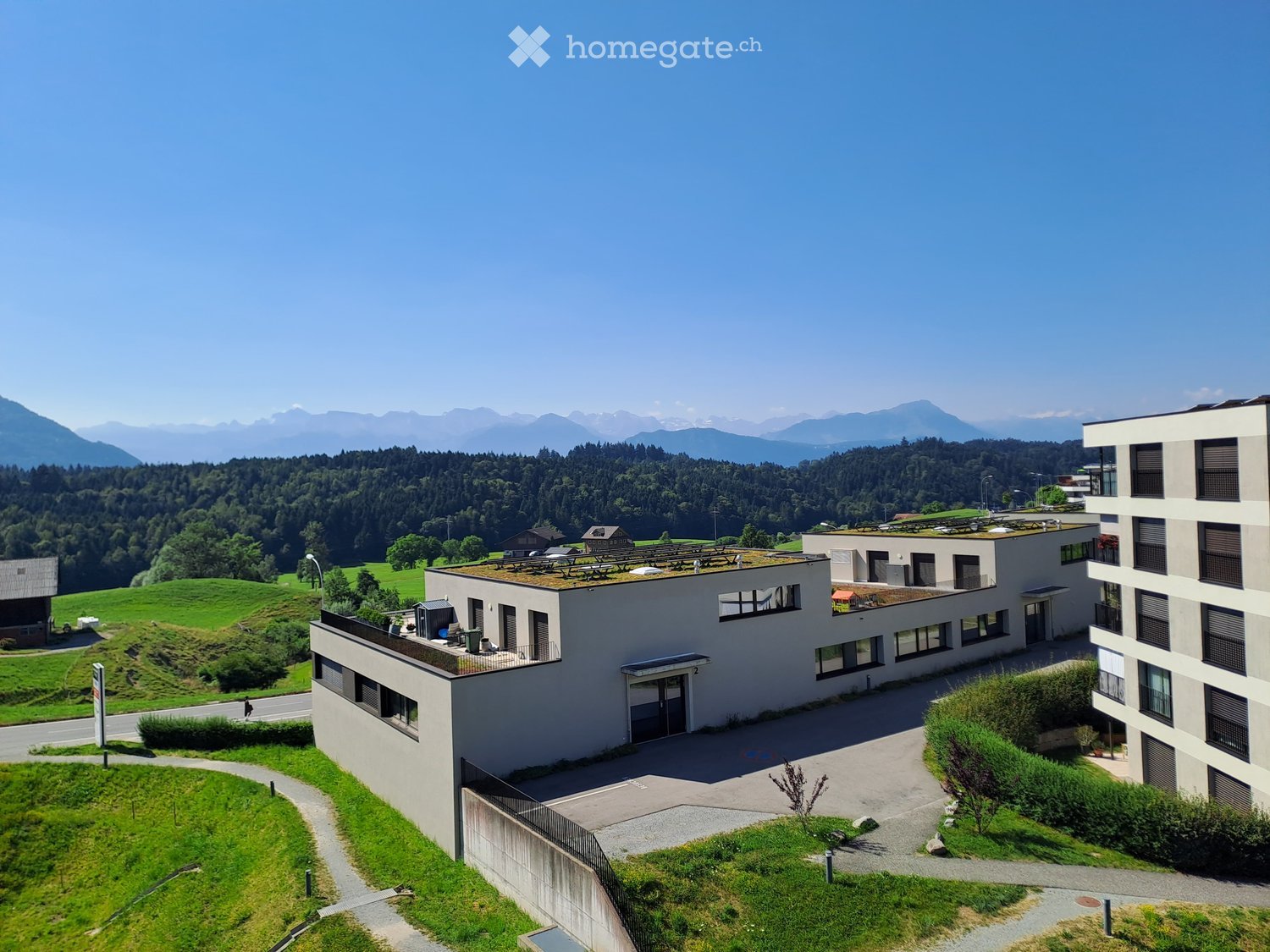 Modern multi-story building with flat roofs and large windows, surrounded by green fields and mountains in the background.