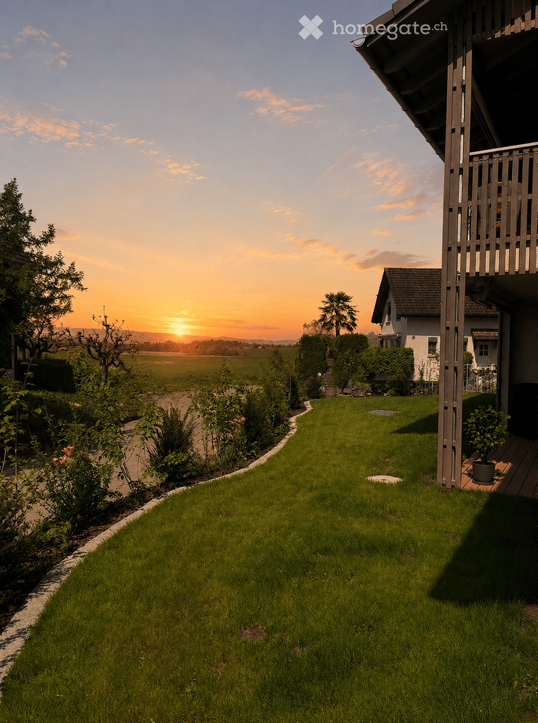 Well-maintained lawn with a curved pathway, surrounded by shrubs and flowers, leading to a house with a balcony and a view of a sunset over a field.
