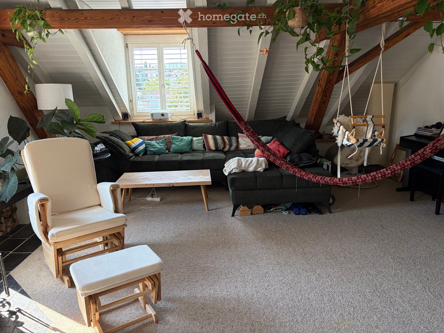 Cozy living room with exposed wooden beams, a green sofa with colorful pillows, a wooden coffee table, a white armchair, a small stool, a hanging hamaca, and a hanging plant.