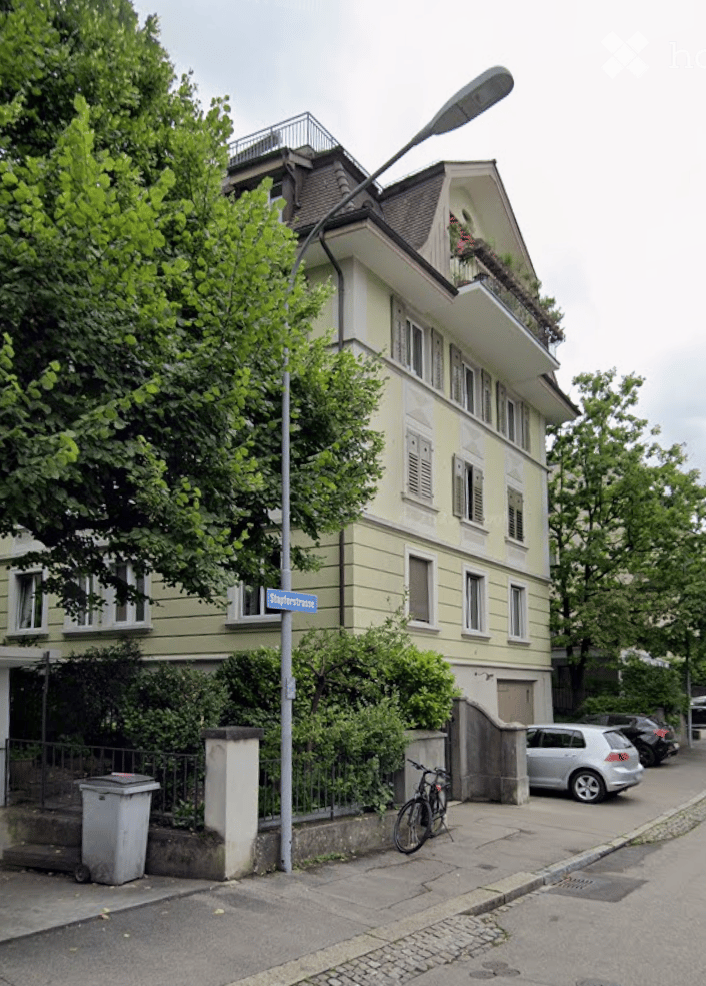 A multi-story residential building with a light yellow facade, multiple windows, and a small balcony on the top floor. The building is surrounded by greenery and has a street sign reading "Schäferbrücke" visible.