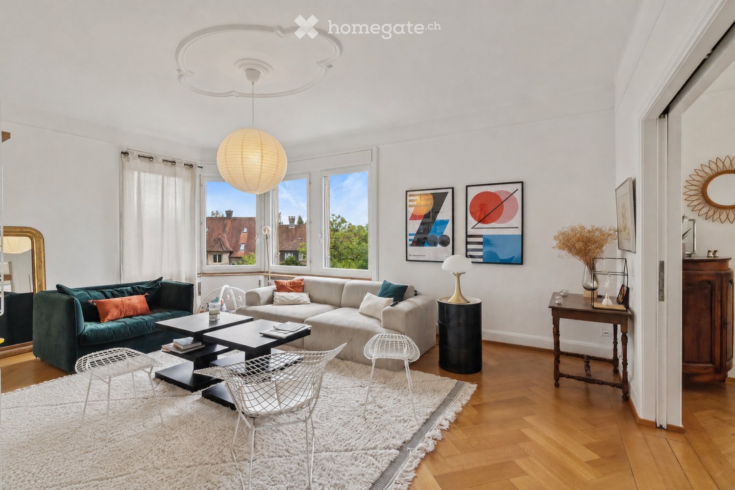 Modern living room with white walls, wooden flooring, large windows, a grey sectional sofa, a green accent sofa, a coffee table, white chairs, a table lamp, and artwork on the walls.