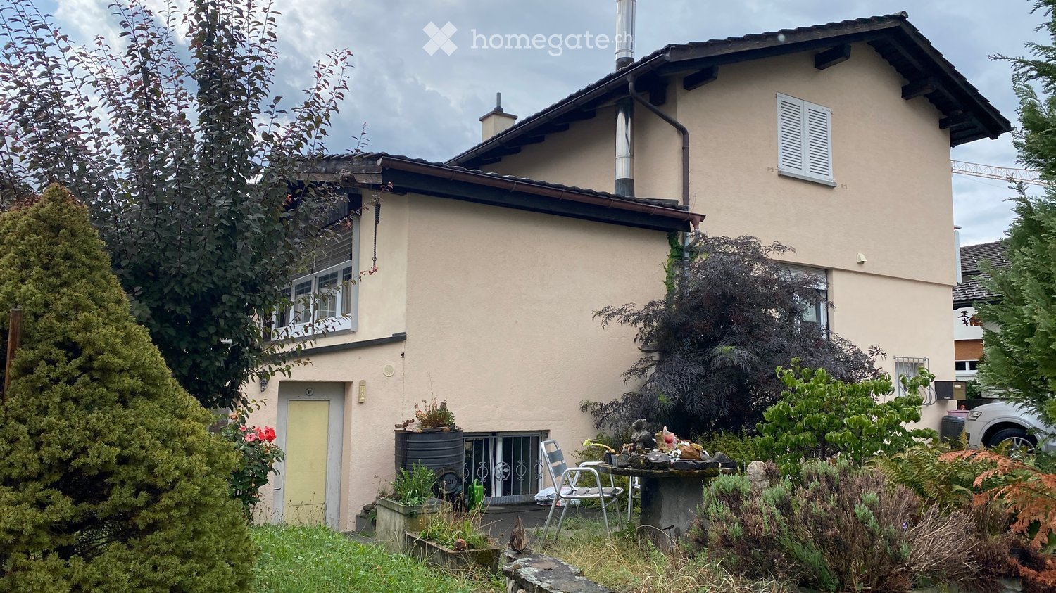 Two-story house with beige exterior, multiple windows, and a small garden area with plants and a table.