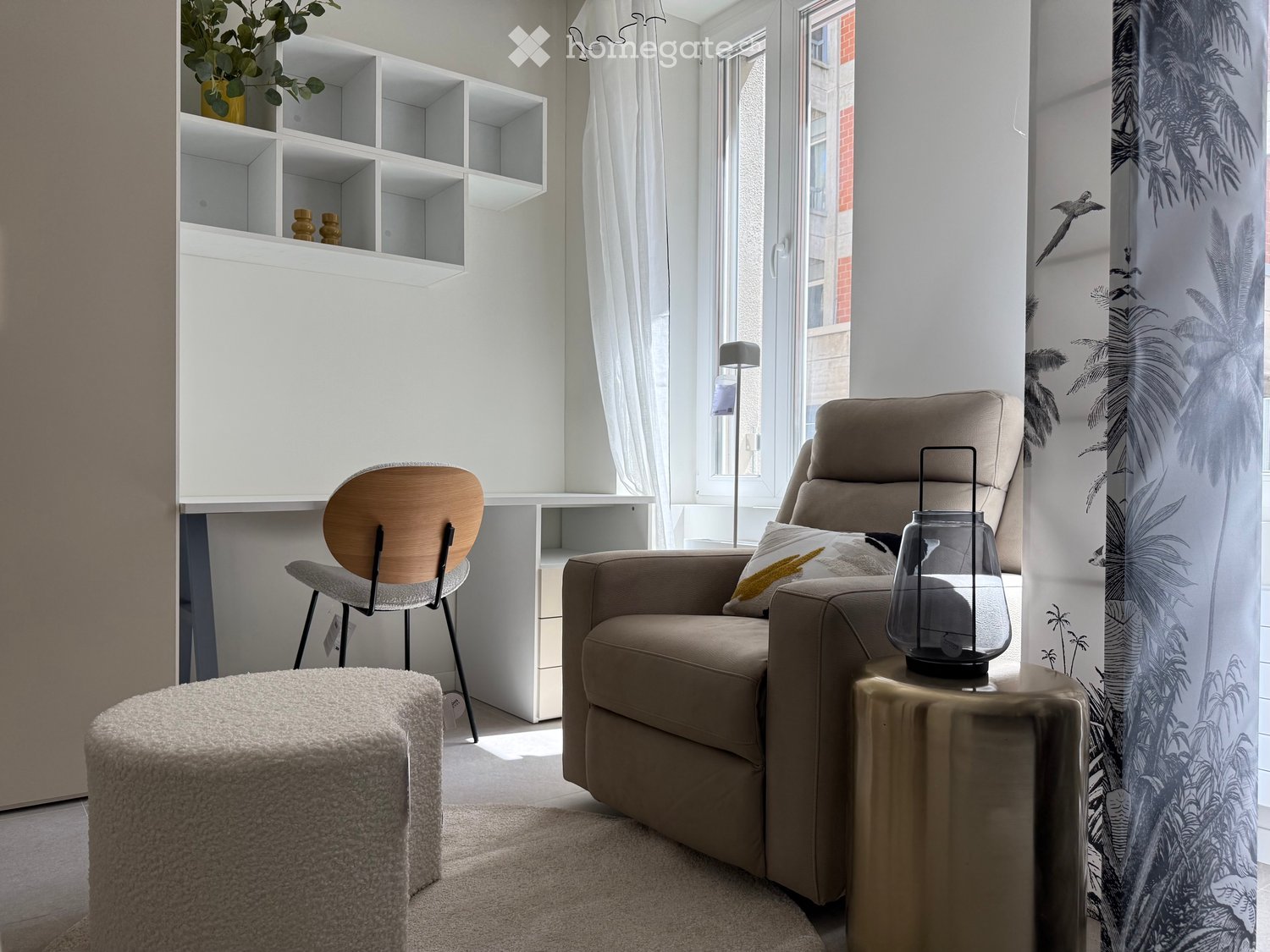 A modern, cozy living room with a beige sofa, wooden chair, round stool, small table with a lantern, white shelving unit, and large windows with sheer curtains.