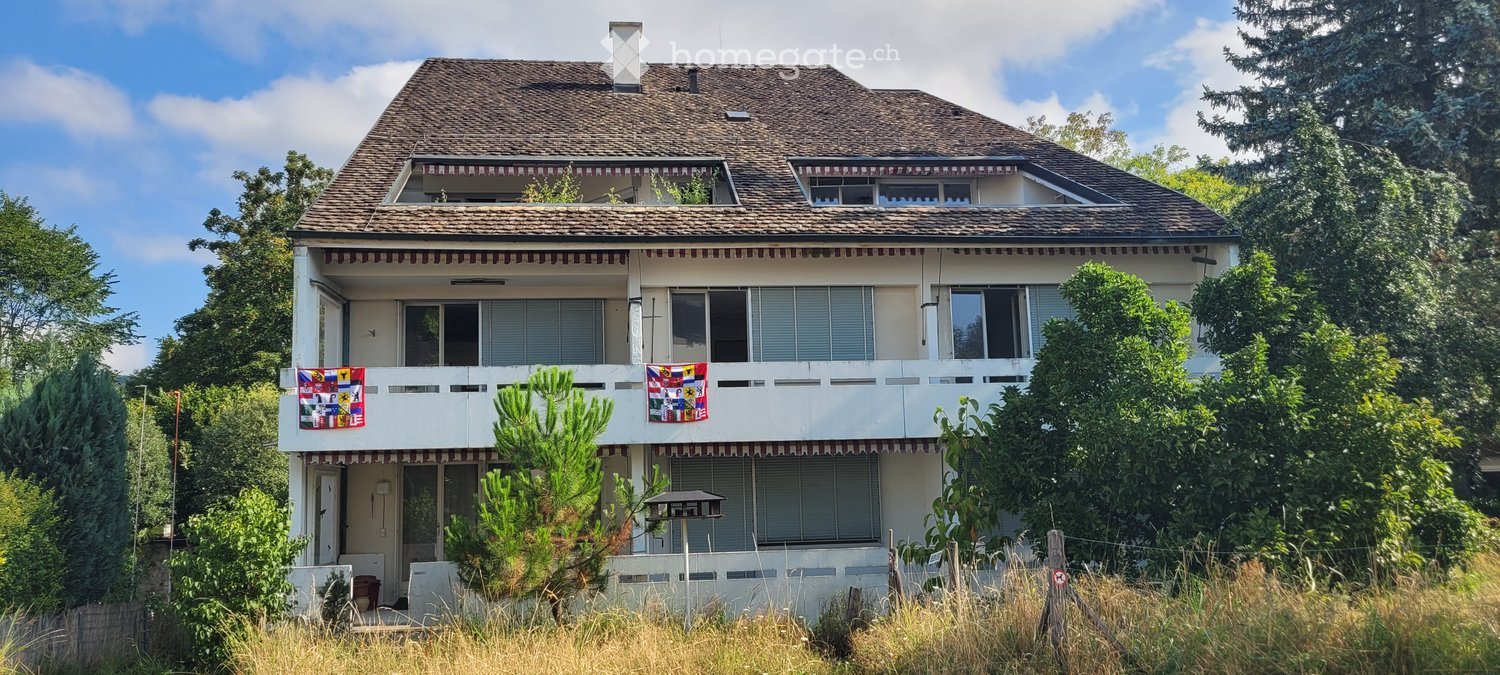 Two-story house with a tiled roof, white exterior, multiple windows, and a balcony with decorative flags.