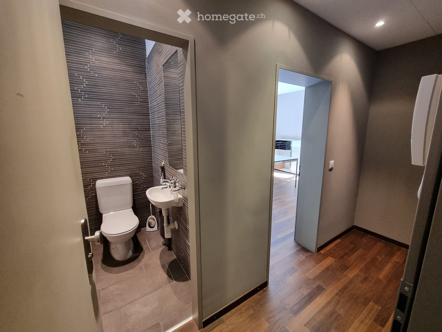 Modern bathroom with a toilet, sink, and faucet, featuring dark tiled walls and a wooden floor visible through an open doorway.