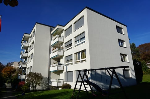 Modern multi-story apartment building with white facade, multiple balconies, and a small playground area in front.