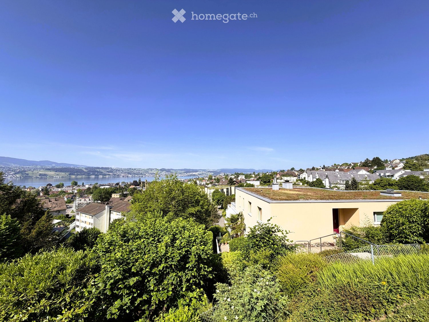 A modern house with a flat roof and a small garden, surrounded by lush greenery and overlooking a scenic lake and town.