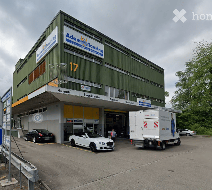 Two-story commercial building with green facade, signage for 'Adam & Touring', garage entrance, and several parked cars including a white convertible and a black sedan.