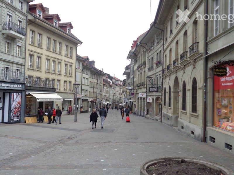 A wide, pedestrian-friendly street lined with multi-story buildings featuring classic European architecture, including balconies and storefronts. People are walking along the street, and there are a few shops visible on the ground level.