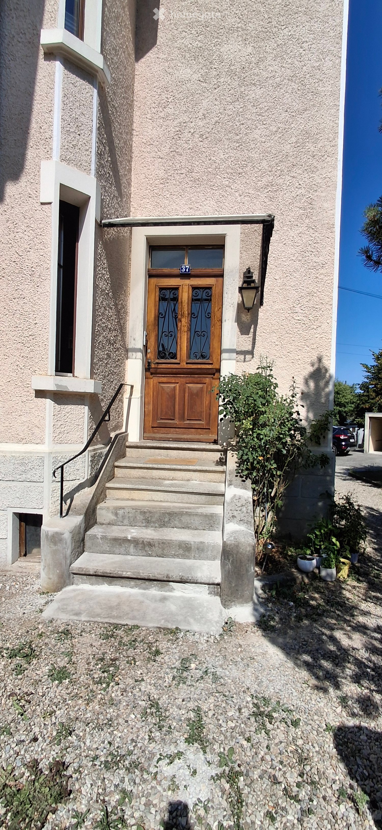 Two-story house with a wooden front door, concrete steps, and small garden area with potted plants.