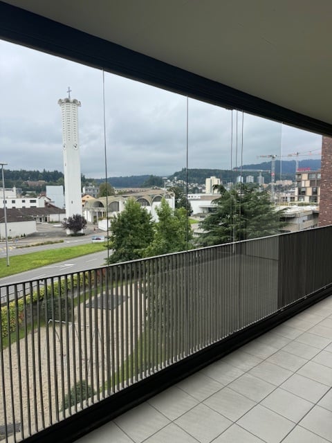 Balcony with modern glass railing, tiled floor, and a view of a cityscape with a prominent tower and surrounding greenery.