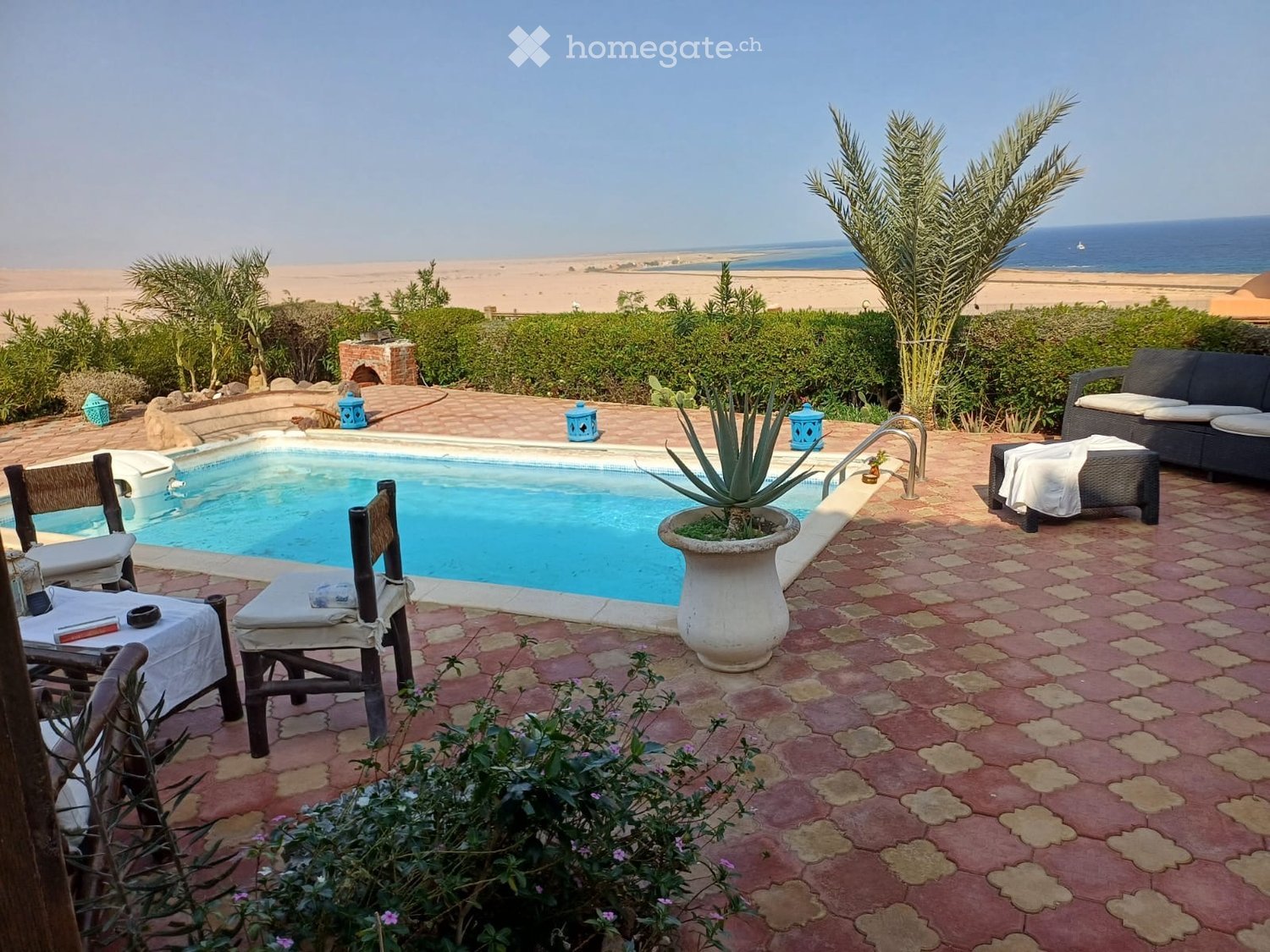 Outdoor area with a swimming pool, surrounded by chairs, a sofa, a table, and potted plants, overlooking the ocean.