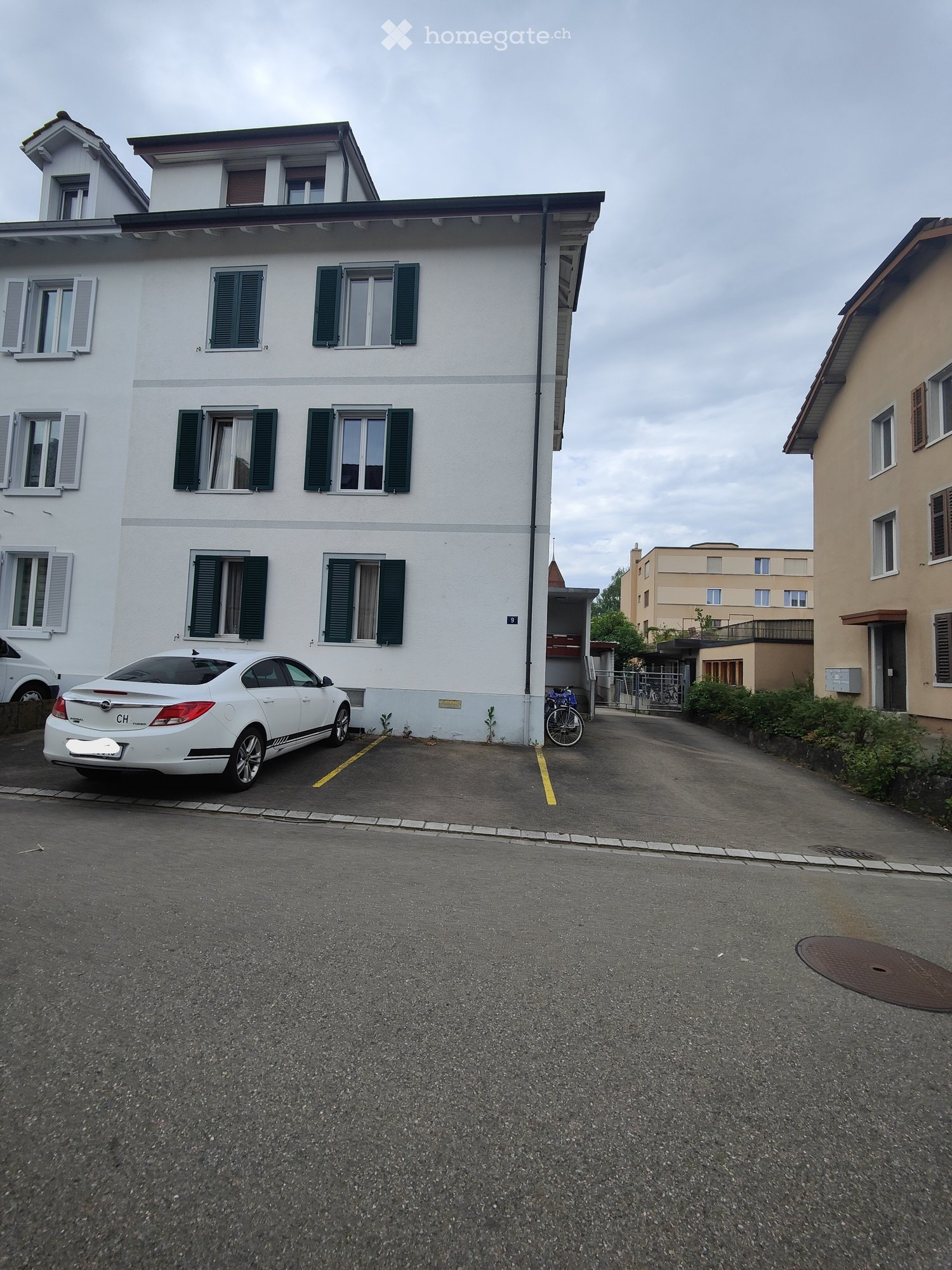 A multi-story residential building with white walls, green shutters, and multiple windows. A white car is parked in front, and a bicycle is near the entrance.