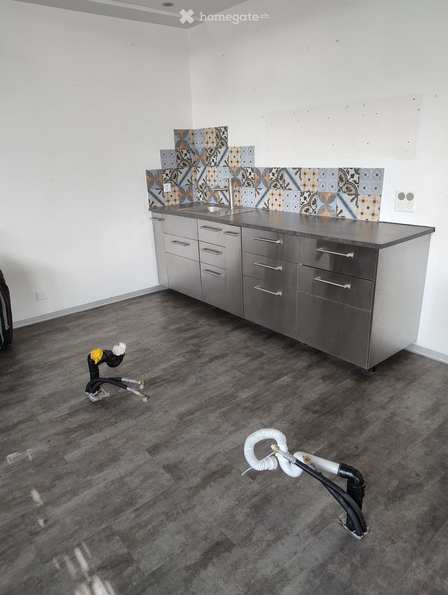 Empty kitchen with stainless steel cabinets, a sink, and a decorative tile backsplash. The floor is made of dark wood planks.