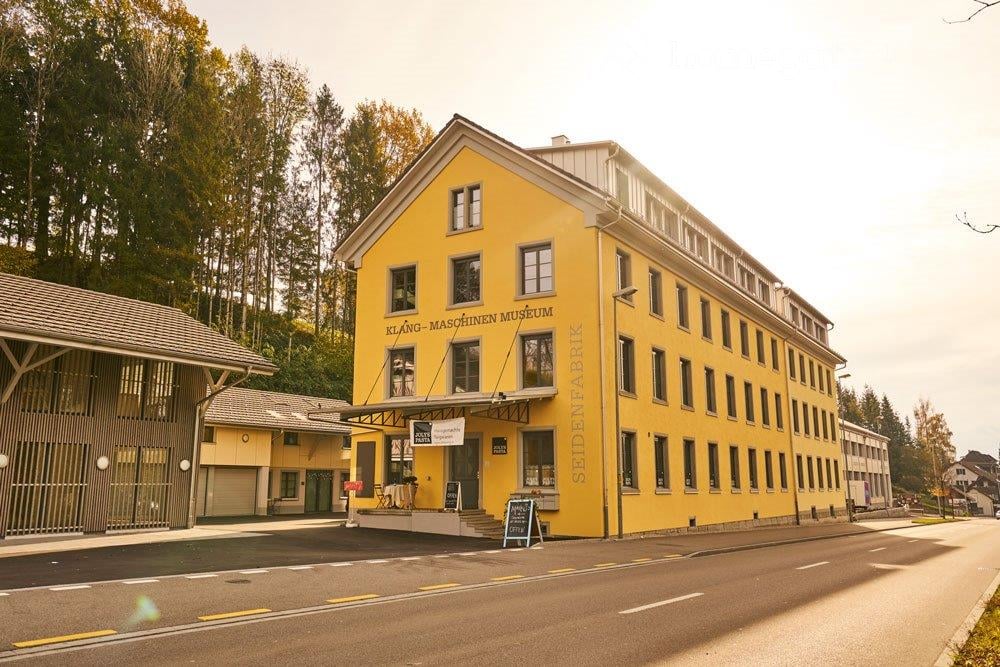 A large, yellow, multi-story building with a flat roof and numerous windows, identified as the Klang-Maschinen Museum. The building has a modern design with a signboard and a small entrance area.