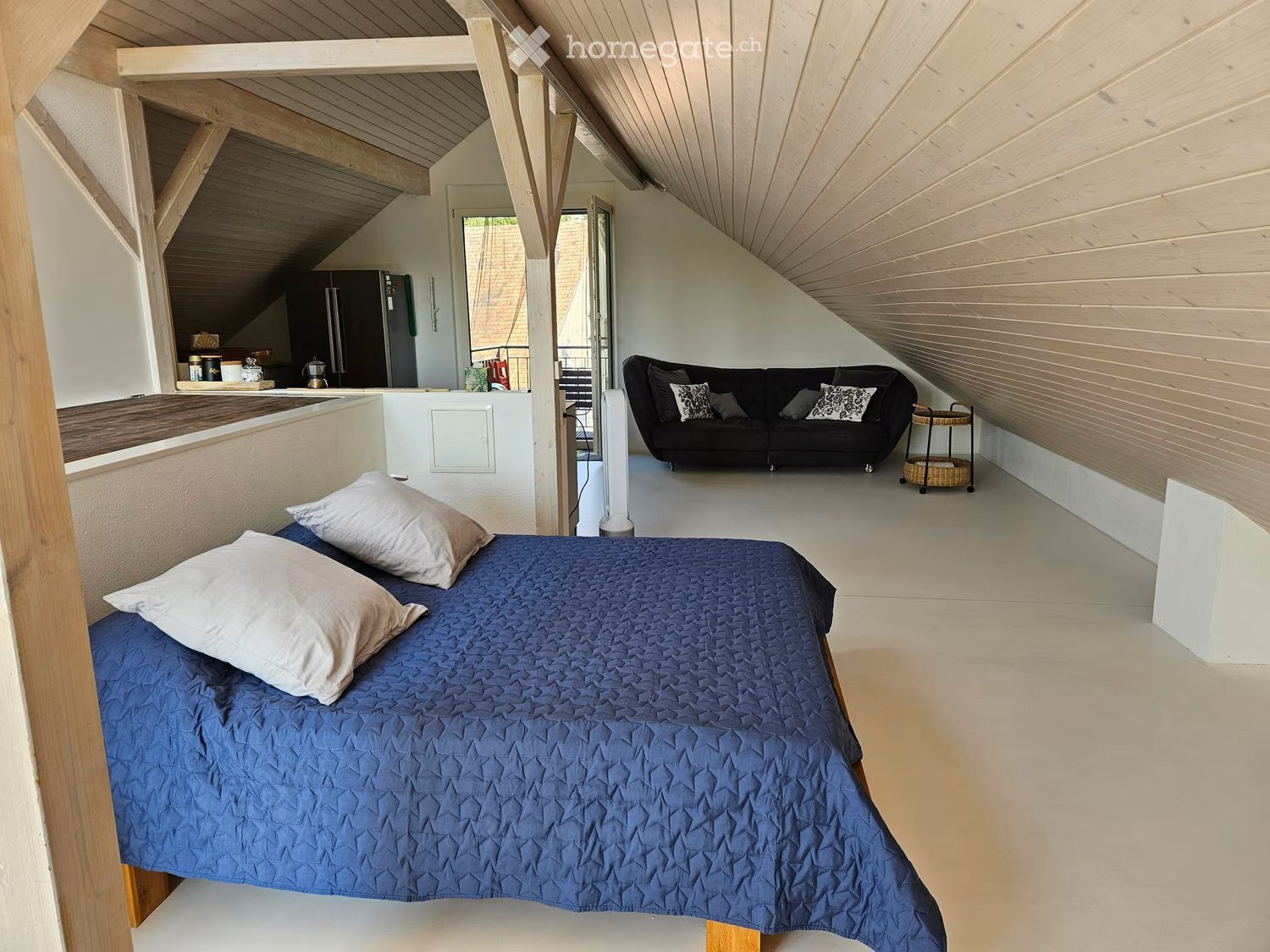 A modern, loft-style bedroom with a blue quilted bed, a black sofa, a small kitchen area with a refrigerator, and a coffee table.