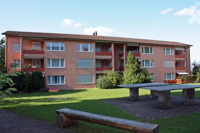 3 story apartment building, brown exterior, multiple windows, balcony, outdoor seating area, green lawn