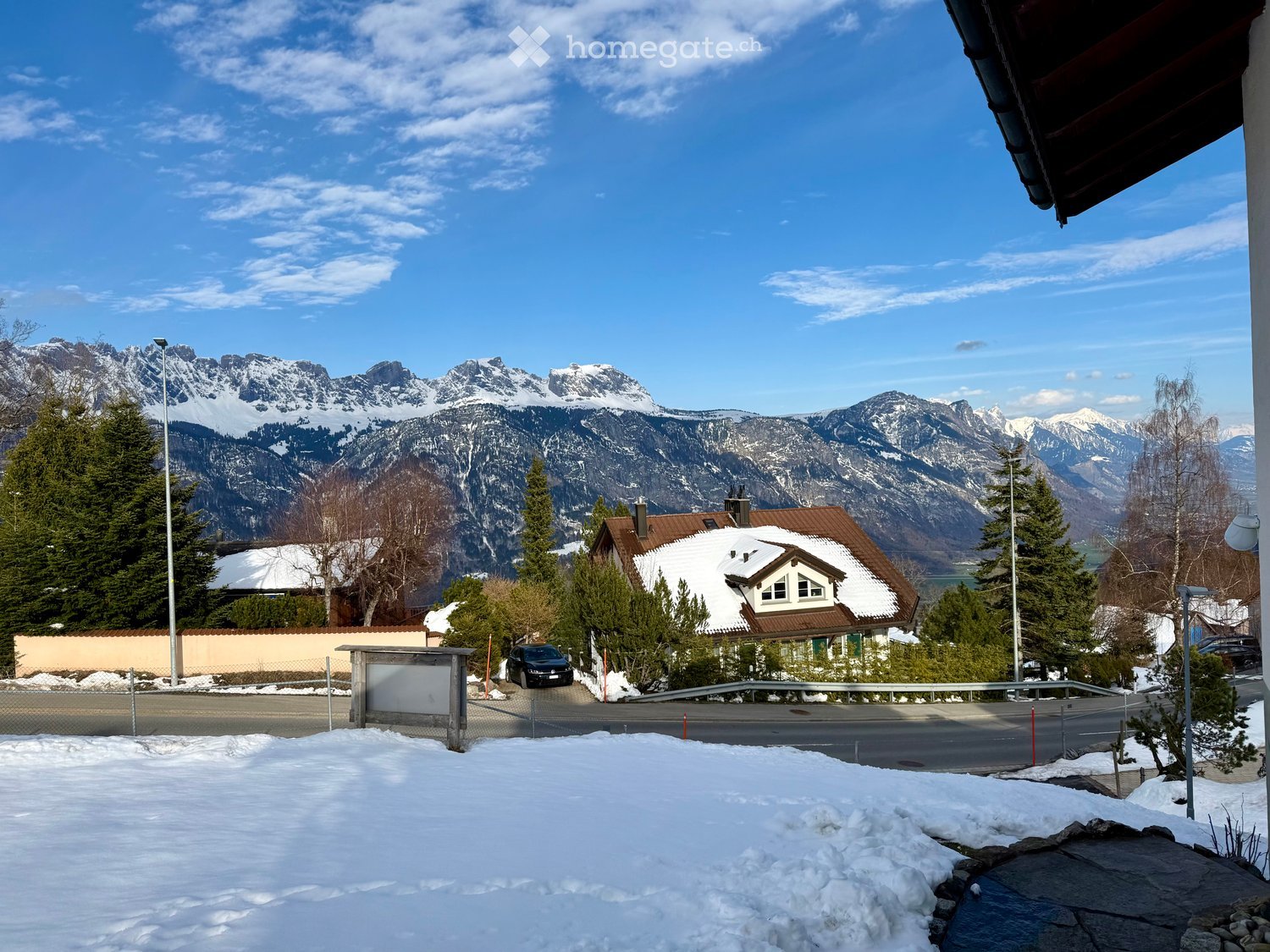 House with brown roof, snow on the roof, car parked in front, surrounded by trees, mountains in the background, snow on the ground