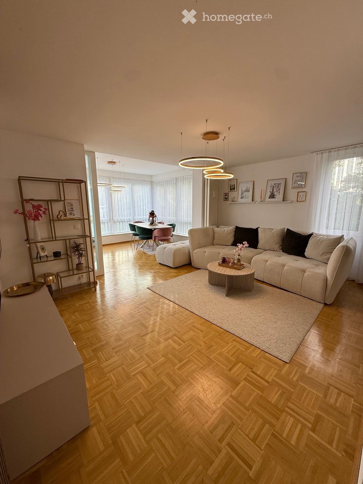 Modern living room with light wooden herringbone flooring, large windows with white curtains, a sectional sofa, coffee table, dining area with chairs and table, and a decorative shelf with vases and picture frames.