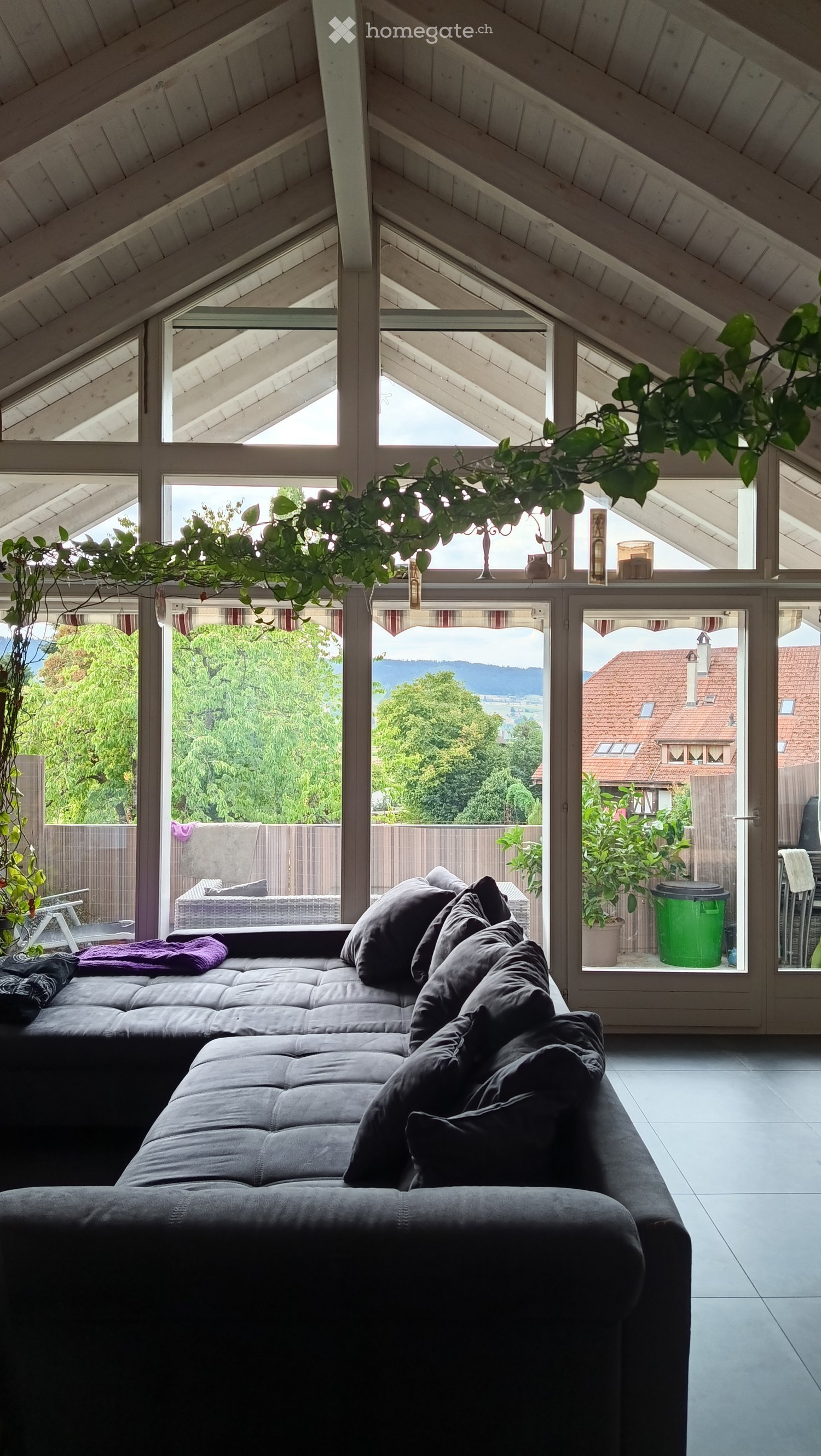 Bright living room with large windows and a view, featuring a dark grey sectional sofa with multiple pillows, a small table with candles, and a hanging plant.