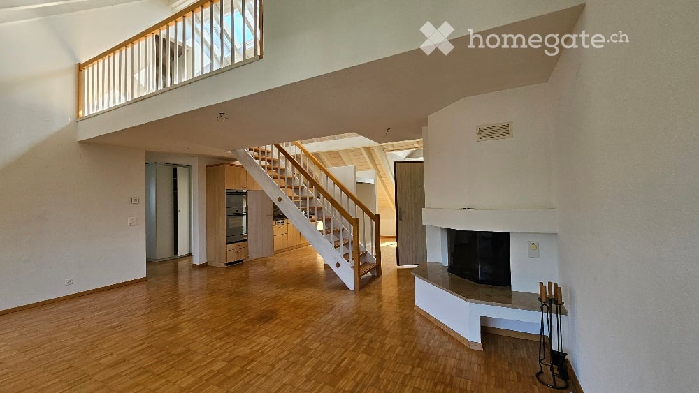 Open-concept living space with wooden flooring, a staircase leading to an upper level, and a fireplace with a white surround. The area includes built-in cabinets and a small kitchen area visible in the background.