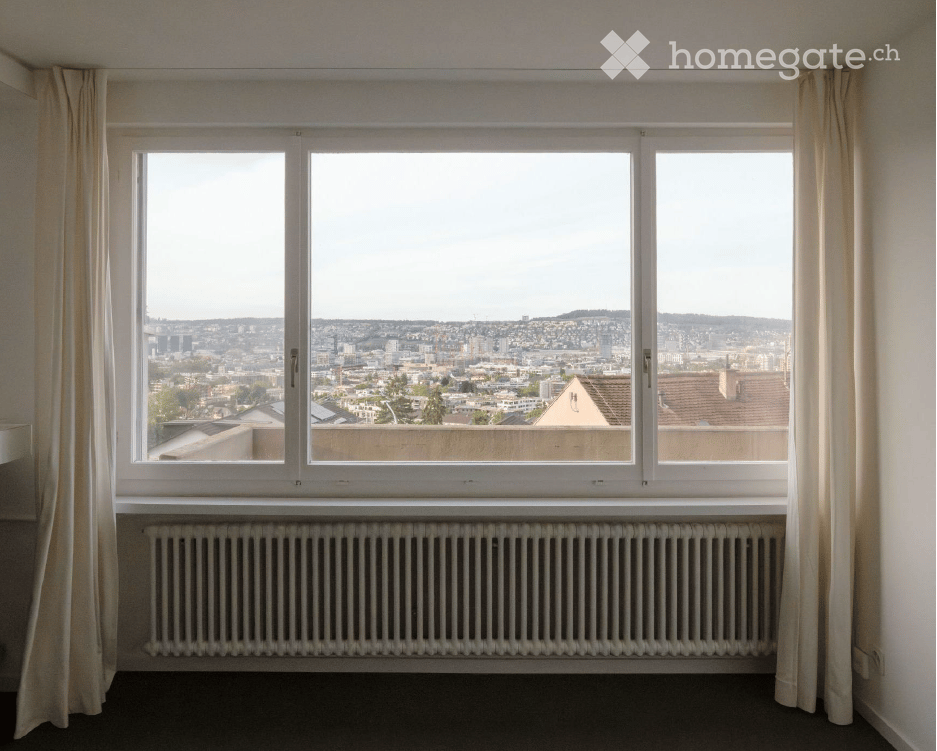 Empty room with large windows offering a cityscape view, white radiator, and light-colored curtains.