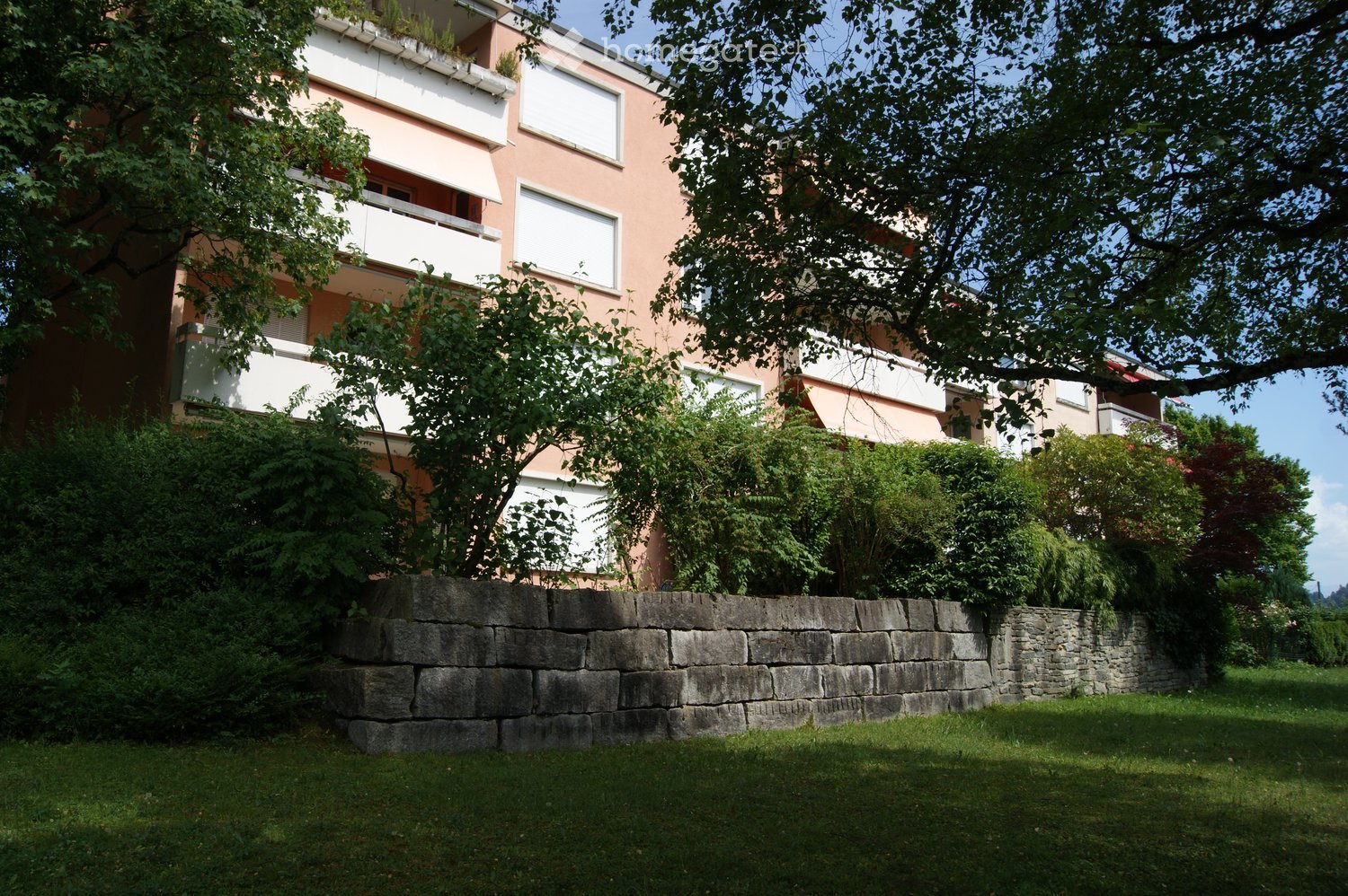 Multi-story residential building with balconies, surrounded by greenery and a stone wall.
