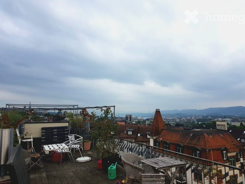 Rooftop terrace with wooden decking, outdoor furniture including chairs and a table, potted plants, and a view of the city skyline and surrounding mountains.