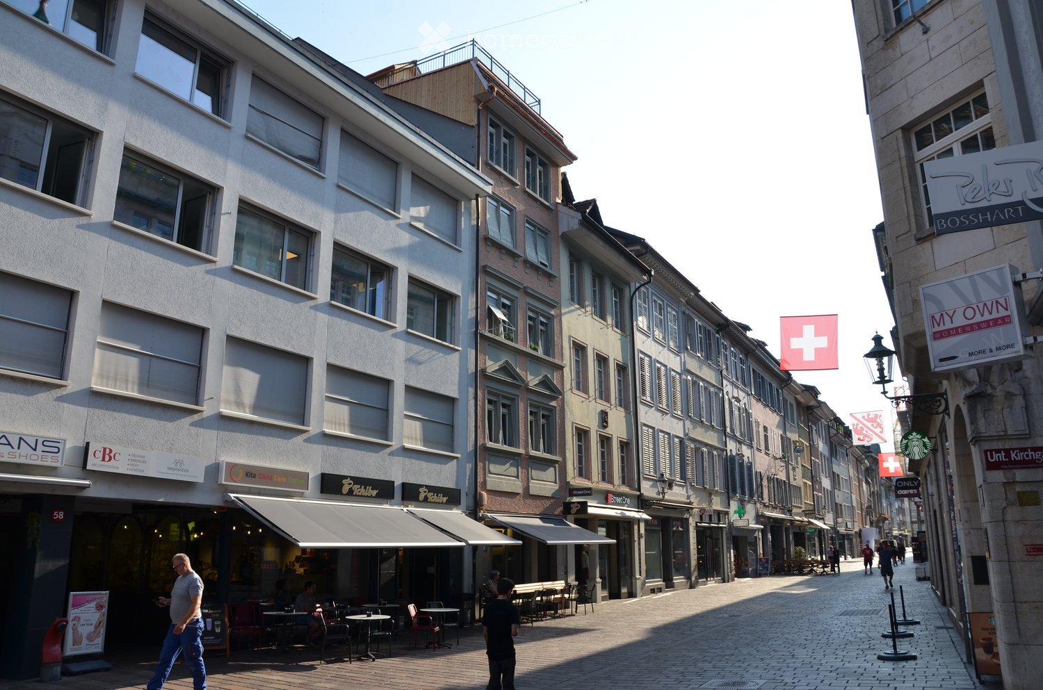 A European-style street lined with multi-story buildings, featuring shops, restaurants, and cafes with outdoor seating areas. The architecture includes classic facades, awnings, and signage for various businesses.