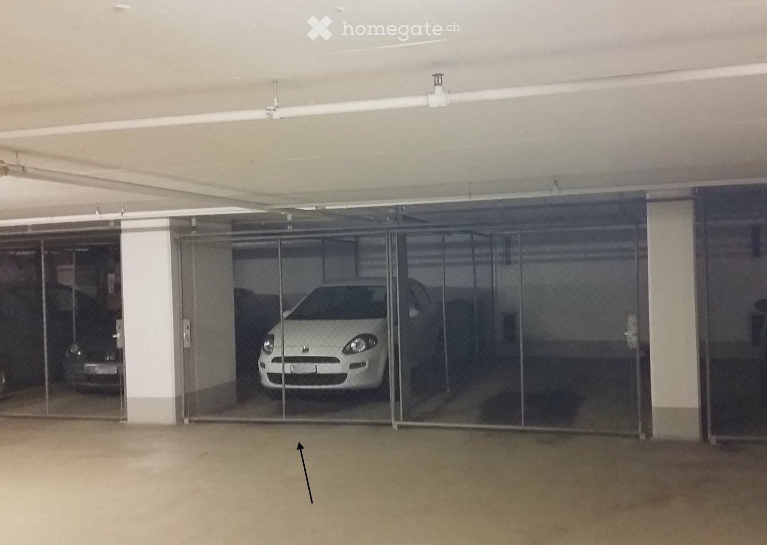 Empty underground parking area with concrete floors and walls, featuring a glass security gate and a white car visible behind it.