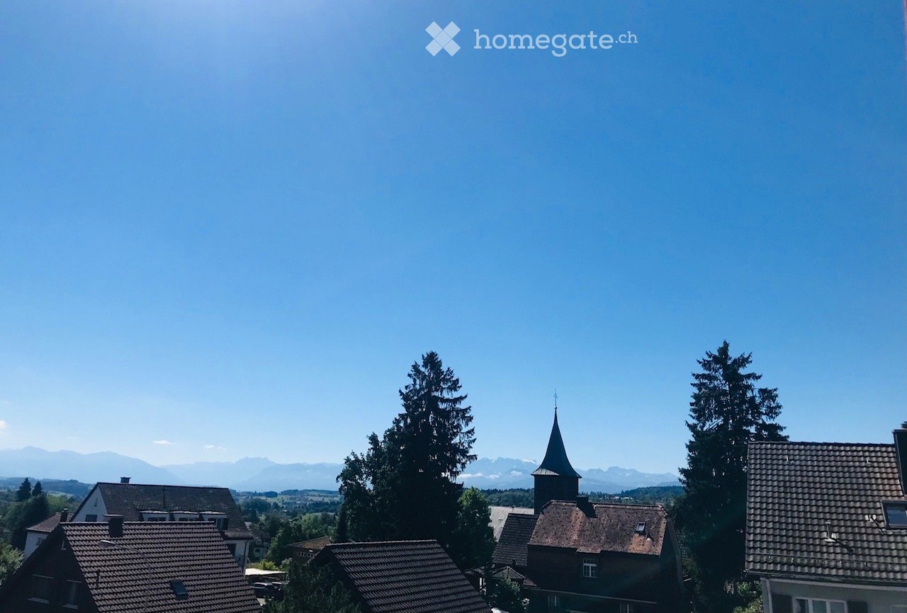 A scenic view of a village with traditional houses, a church tower, and surrounding mountains under a clear blue sky.