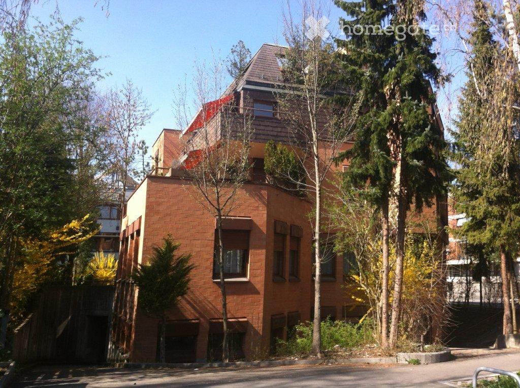 Modern two-story brick building with large windows and a sloped roof, surrounded by trees and greenery.
