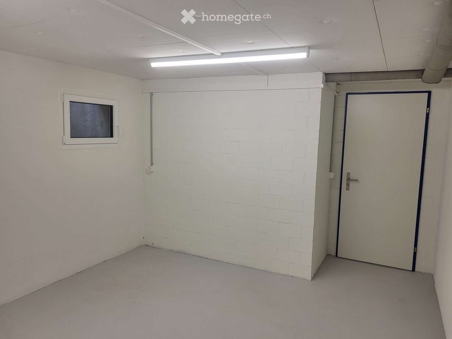 Empty room with white brick walls, a single window, and a door. The space is unfurnished with a smooth concrete floor and a white ceiling with recessed lighting.