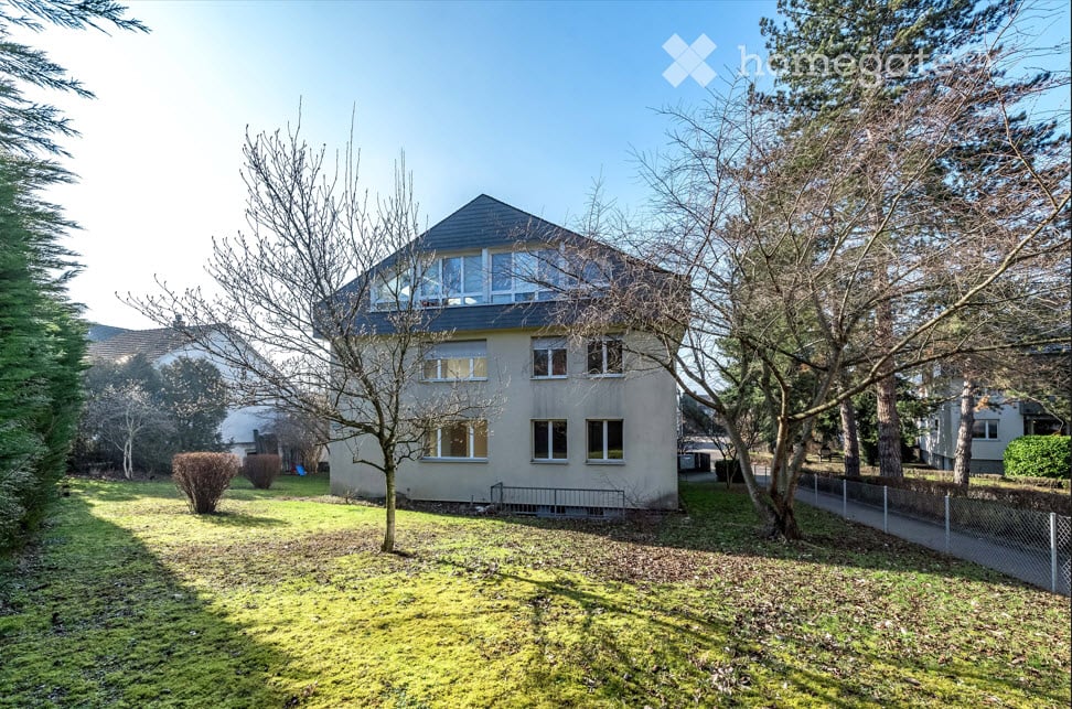Two-story house with a flat roof and large windows, surrounded by trees and a grassy lawn.
