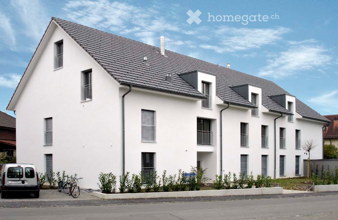 Modern two-story white building with grey roof, multiple windows, balconies, and small garden area in front.