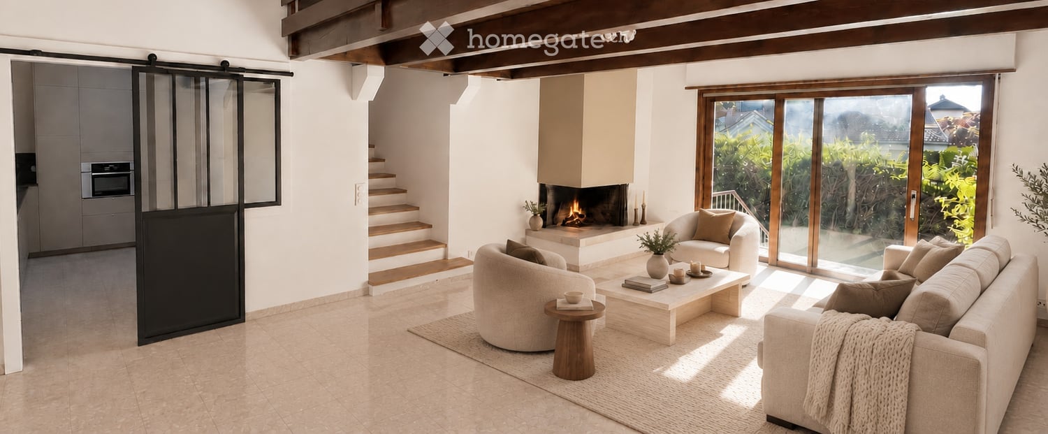 Modern living room with a fireplace, large windows, and comfortable seating including a sofa, armchair, and ottoman. There is a coffee table, a small wooden stool, and an oven visible in the background.