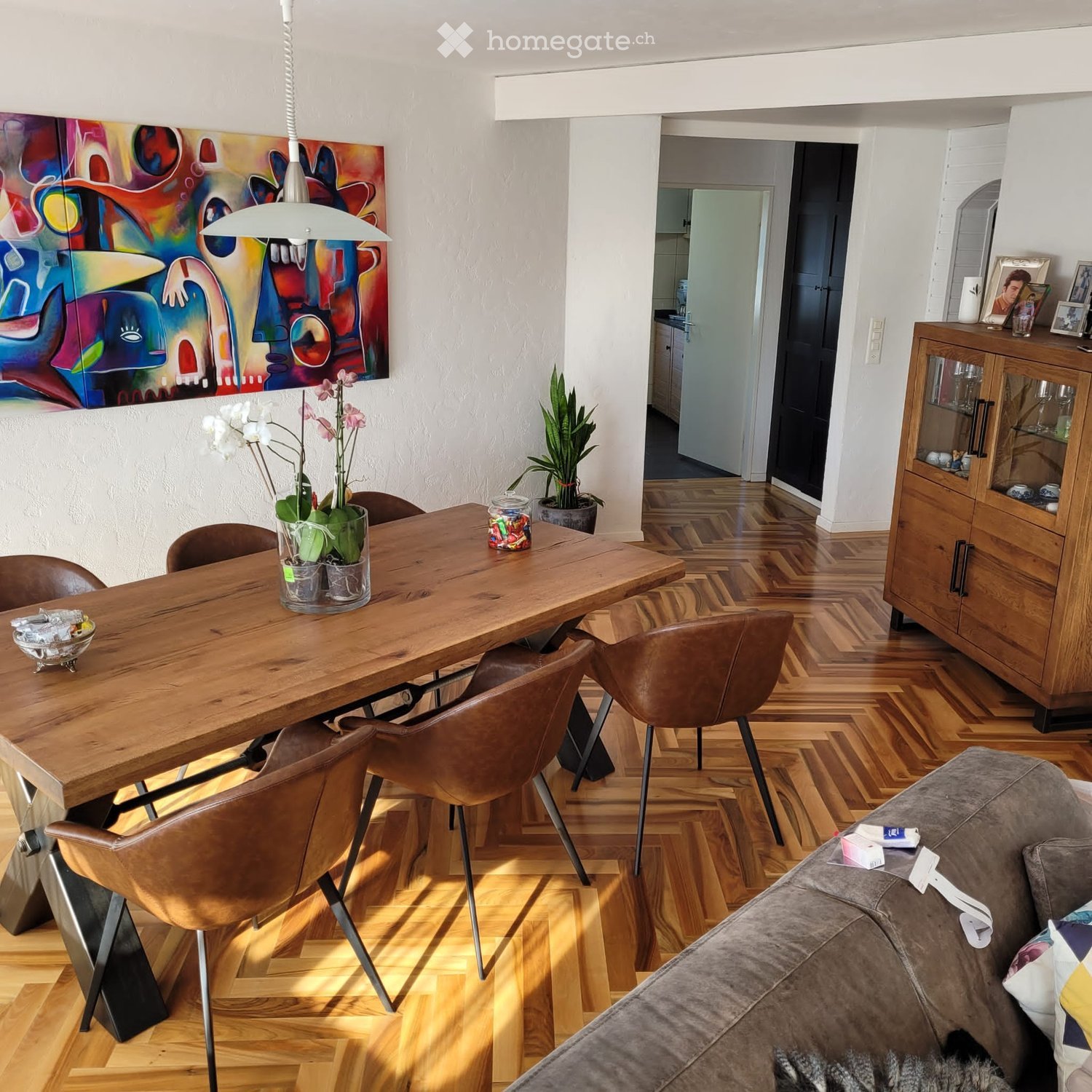 Modern living and dining area with a grey sofa, wooden dining table, leather chairs, a wooden cabinet, and a colorful abstract painting on the wall.
