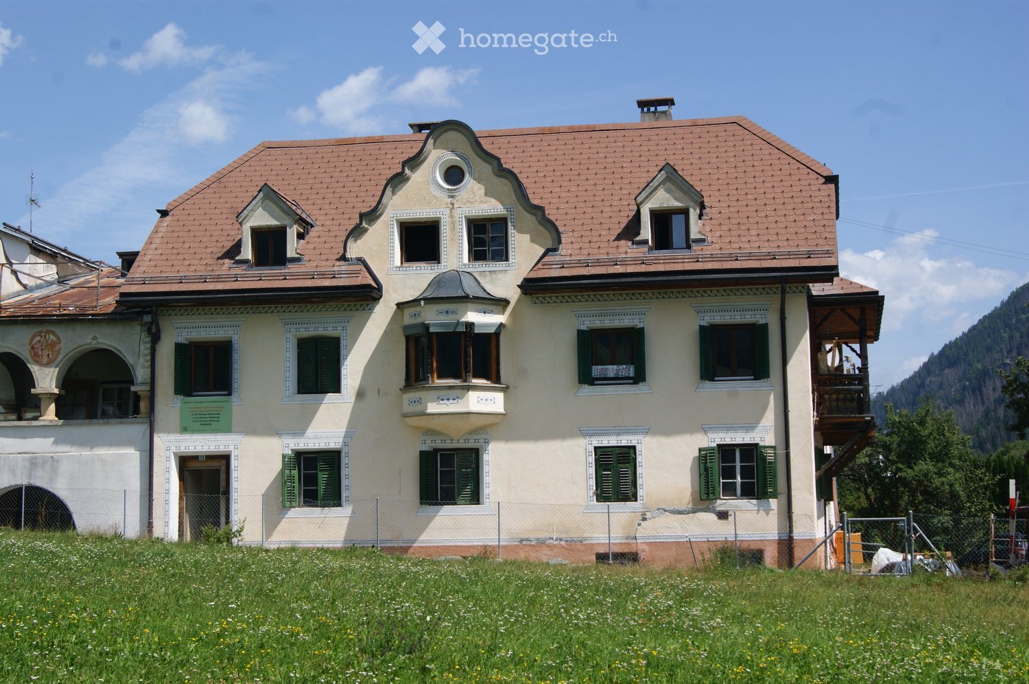 A two-story house with a red-tiled roof, multiple windows with green shutters, and a balcony on the right side. The house is surrounded by a grassy area with wildflowers.