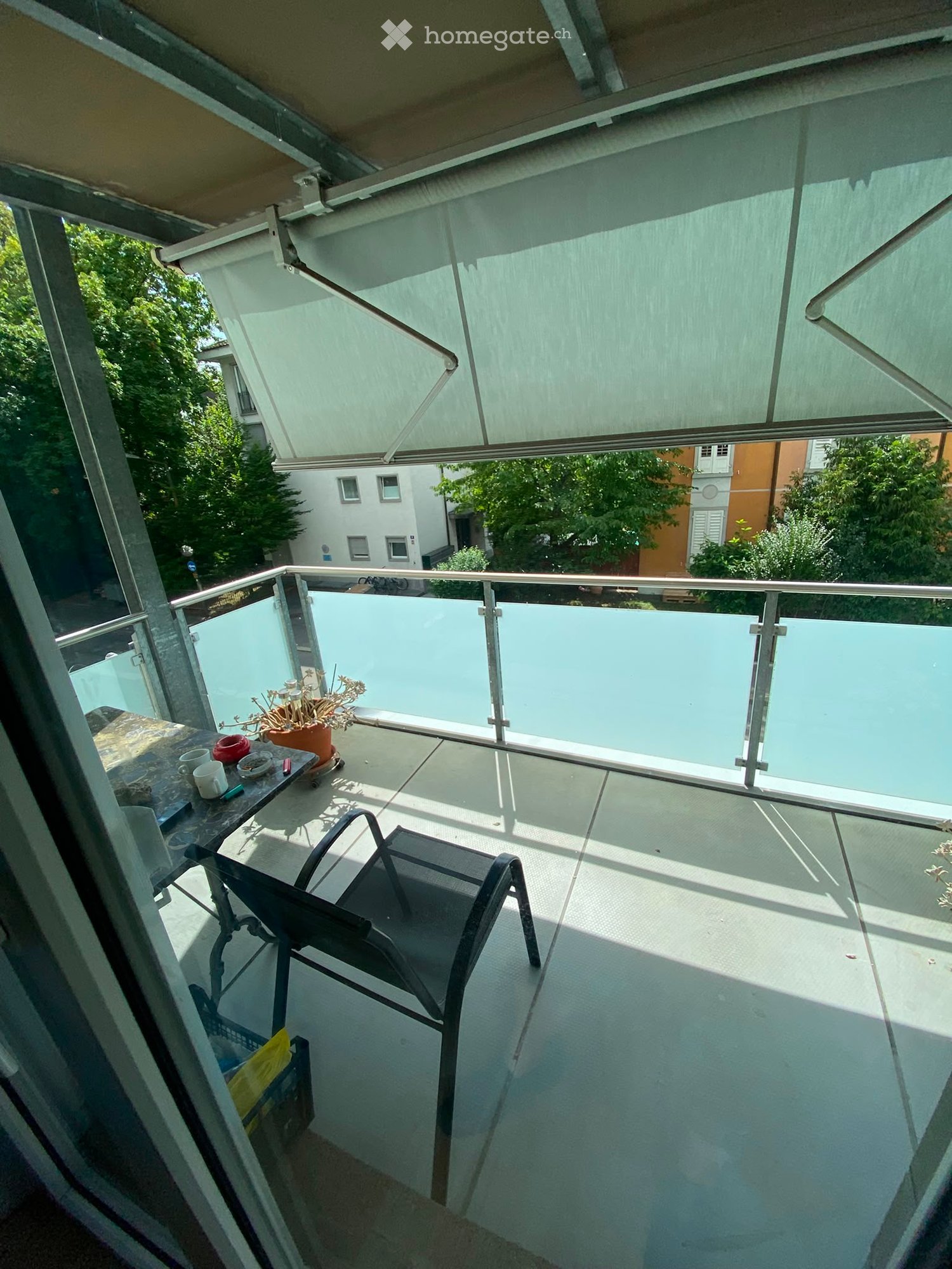 Balcony with a glass railing, a small table with cups and a potted plant, and a folding chair.