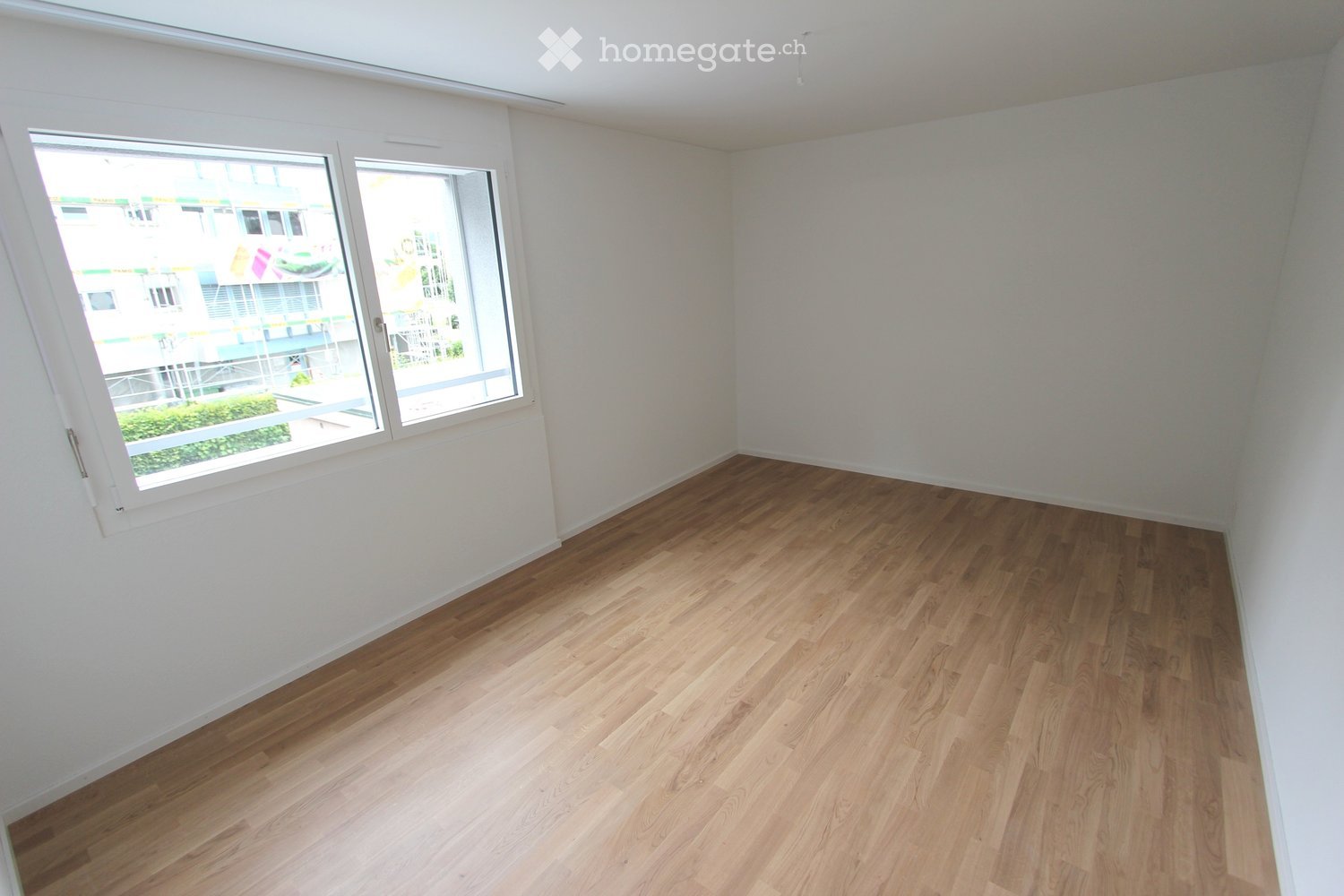 Empty room with wooden flooring and white walls, featuring large windows with a view of an outdoor area.
