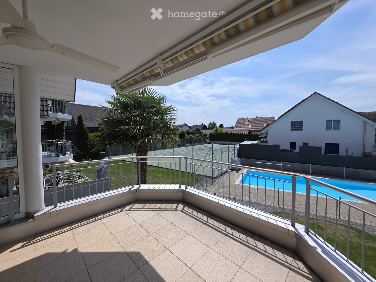 Balcony with tiled floor, metal railing, retractable awning, overlooking a swimming pool, tennis court, and surrounding houses.