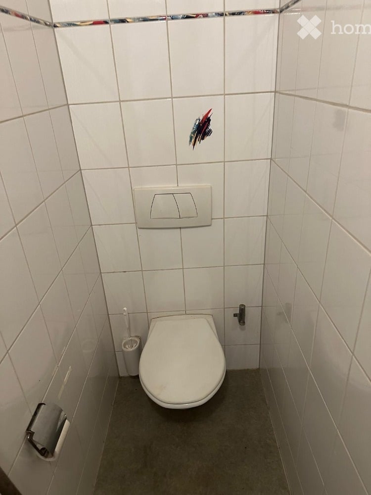 A small bathroom with white tiled walls and a grey floor. It features a toilet, a toilet paper holder, and a toilet brush holder.