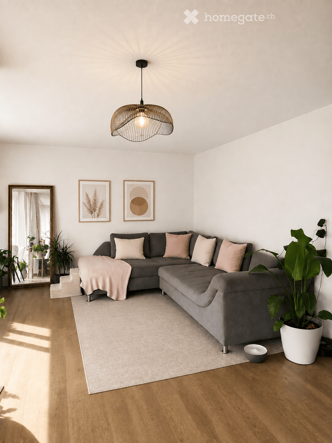 Modern living room with grey sectional sofa, throw blanket, large potted plant, mirror, picture frames, and a hanging pendant light.