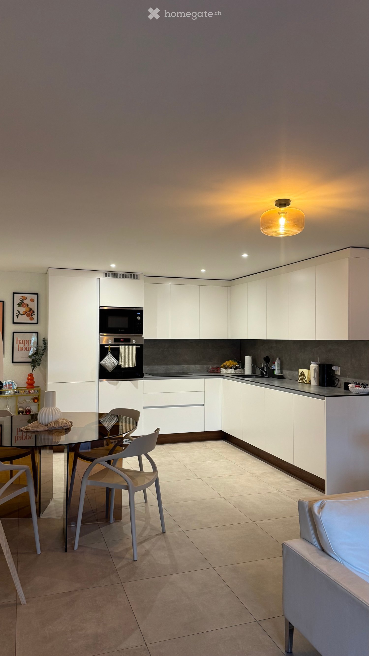 Modern kitchen with white cabinets, glass dining table, chairs, and various appliances including an oven, microwave, and coffee maker. The space features a sleek design with a tiled floor and a pendant light.