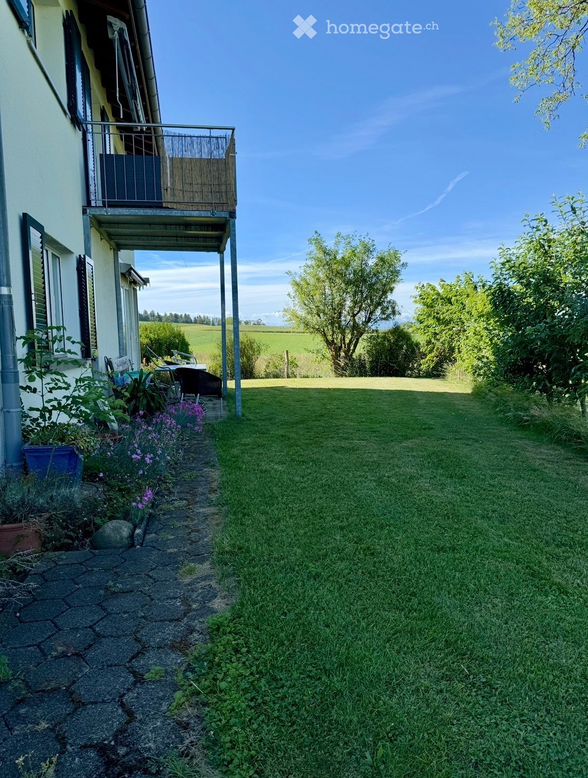 Garden area with a stone pathway leading to a grassy lawn, surrounded by trees and a house with a balcony.