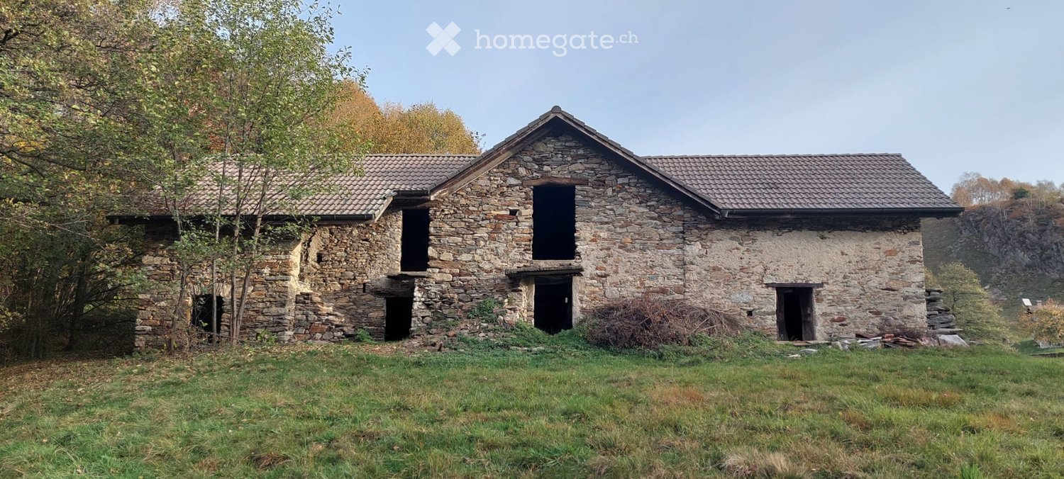 A rustic stone building with a tiled roof, multiple windows, and doors, surrounded by grass and trees.